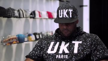 La casquette noire à taches blanches UTK Unkut de Booba dans la vidéo "Derrière la plume de Booba, le bitume d'Élie Yaffa"
