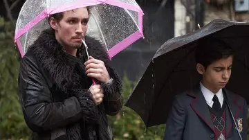 Abrigo negro con cuello de piel usado por Klaus (Robert Sheehan) como se ve en el vestuario de la serie de televisión The Umbrella Academy (Temporada 1 Episodio 1)