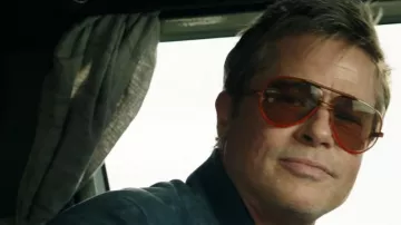 Close-up film still of Brad Pitt inside a car, wearing a navy blue suede bomber jacket and vintage double-bridge aviator sunglasses with clear orange lenses. used by Sonny Hayes (Brad Pitt) in F1